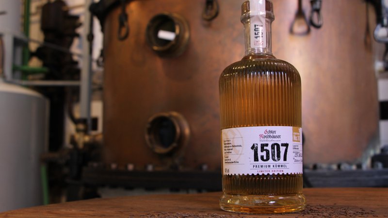 Eine Flasche Echter Nordhäuser Kümmel auf einem dunklen Holztisch vor einer Brennblase aus Kupfer