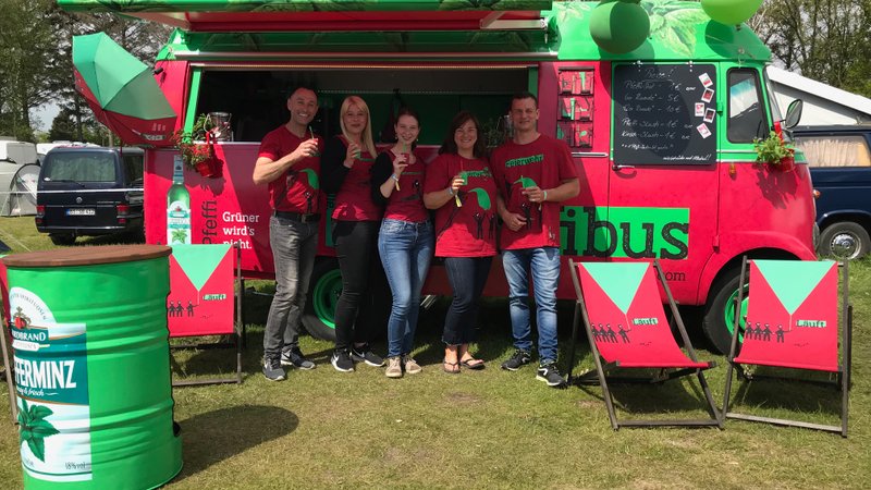 Fünf Personen in roten T-Shirts stehen vor einem bunten Foodtruck, während sie Getränke in der Hand halten und lächeln.