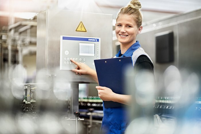 A smiling woman in work clothes operates a control panel in a modern factory environment.