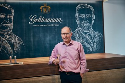Ein Mann in einem rosa Hemd hält ein Glas Sekt und lächelt vor einer Wand mit einer Zeichnung und dem Schriftzug 'Geldermann'.