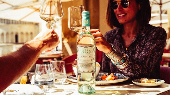 Ein Mann und eine Frau stoßen mit Weingläsern an, während eine Flasche Doppio Passo Moscato auf dem Tisch steht.
