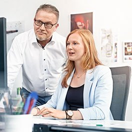 Ein Mann und eine Frau sitzen an einem Schreibtisch, während sie gemeinsam auf einen Computerbildschirm schauen.