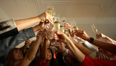 A group of people raise their champagne glasses in a celebratory toast. The focus is on the hands and glasses, with various people smiling in the background under warm lighting.
