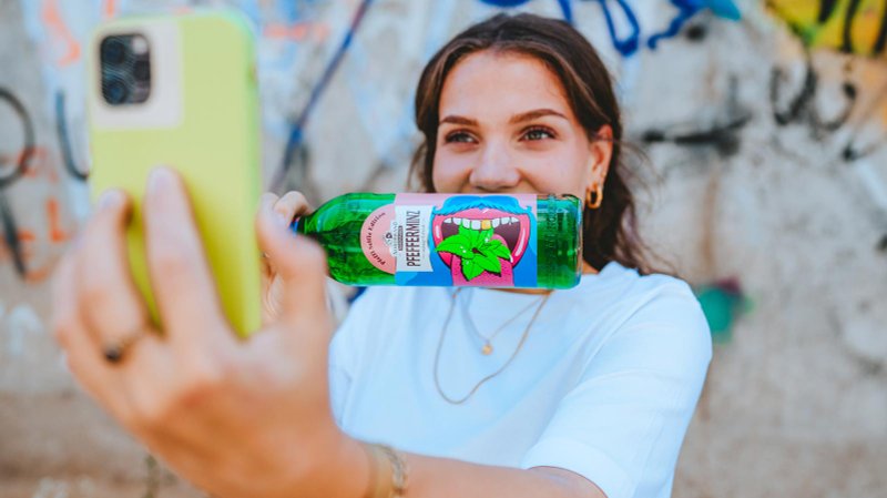 Mit einem fröhlichen Ausdruck hält die Frau eine Flasche Pfeffi in der Hand, während sie ein Selfie vor einer bunten Wand macht.