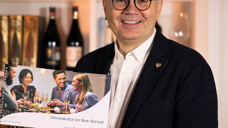 Ein lächelnder Mann hält ein Buch mit dem Titel 'Genusskultur im New Normal' vor einer Bar mit Weinflaschen im Hintergrund.