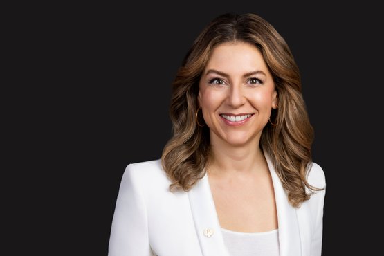 Silvia Wiesner: A smiling woman with long, curly hair wears a white blazer and gold earrings against a dark background.