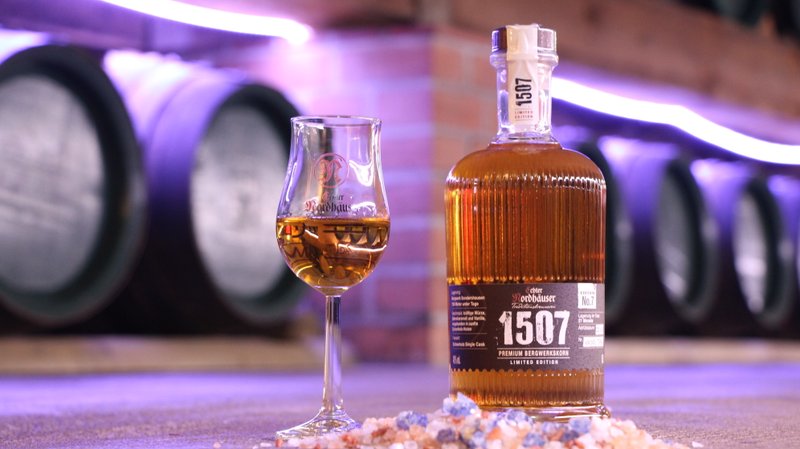 Eine Whiskyflasche und ein Glas auf einem Tisch mit Salzsteinen, im Hintergrund Fässer.