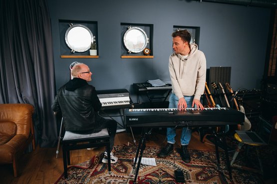 Zwei Musiker in einem stilvollen Raum, einer spielt Klavier, während der andere aufmerksam zuschaut und lächelt.