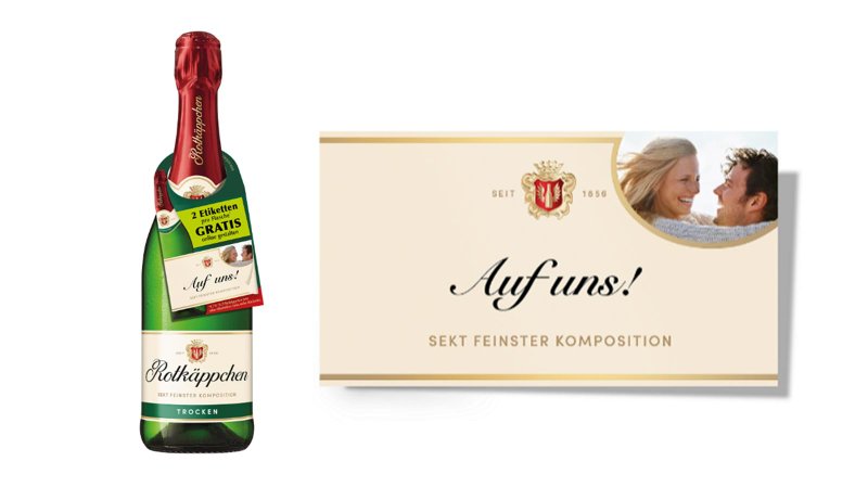 Das Etikett der Sektflasche zeigt den Schriftzug 'Auf uns!' und ein Bild eines glücklichen Paares, das sich anlächelt.