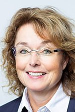 Eine lächelnde Frau mit lockigem, braunem Haar trägt eine Brille und einen dunklen Blazer vor einem hellen Hintergrund.