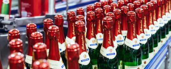 Eine Reihe von Rotkäppchen Mumm Sektflaschen mit roten Kapseln steht ordentlich auf einem Förderband in einer Produktionsstätte.