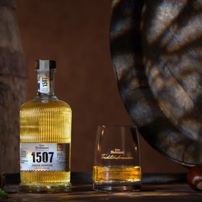 Flasche „1507“ mit goldenem Destillat neben Glas auf Holztisch, im Hintergrund Holzfässer.