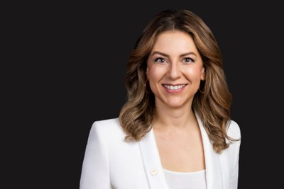 Silvia Wiesner: A smiling woman with long, curly hair wears a white blazer and gold earrings against a dark background.