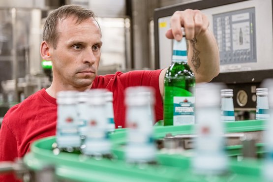 Nordbrand Nordhausen: In a factory environment, a man works holding a green bottle while monitoring bottles on a conveyor belt.