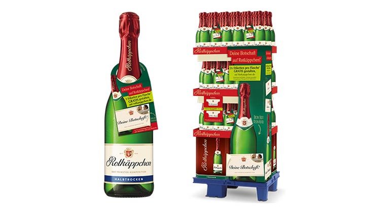 Sag es mit Rotkäppchen: Eine Flasche Rotkäppchen Sekt steht neben einem Verkaufsdisplay mit mehreren Flaschen, das festlich dekoriert ist.