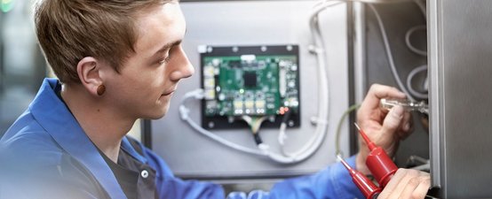 Ein junger Techniker arbeitet konzentriert an einem elektrischen Gerät, während er mit einem Schraubendreher eine Verbindung überprüft.