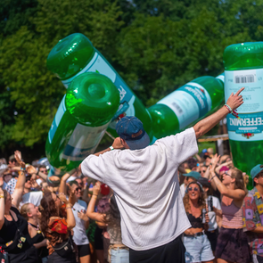 Ein begeisterter Sänger steht vor einer jubelnden Menschenmenge, während riesige grüne Pfeffi-Flaschen durch die Menge getragen werden.