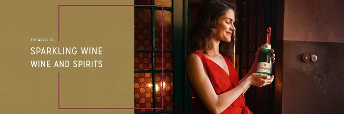 The woman stands in front of a window and holds up a bottle of Rotkäppchen sparkling wine with a smile.