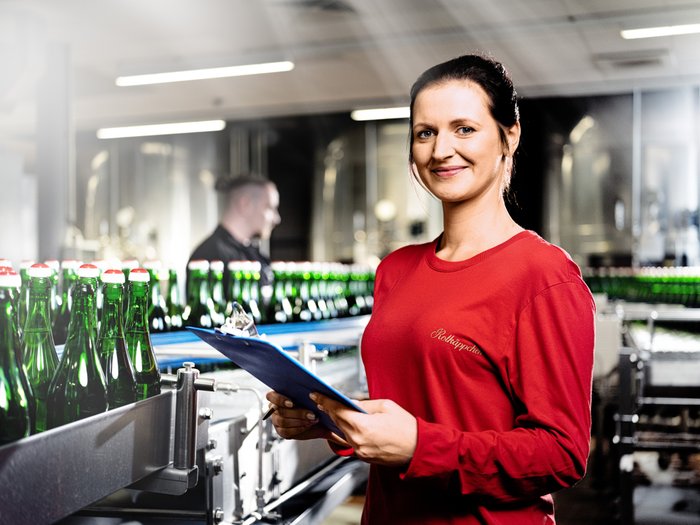 An employee in a red shirt smiles while working in a bottle production line and taking notes.