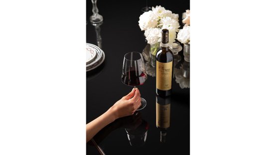 Ein elegantes Weinglas wird von einer Hand gehalten, während eine Flasche Wein und frische Blumen auf dem Tisch stehen.