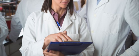 Eine Frau in einem weißen Laborkittel hält einen Clip-Ordner und zeigt auf etwas, während sie mit einem Kollegen spricht.