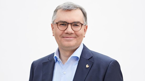 Ein lächelnder Mann mit Brille trägt einen dunklen Anzug und ein hellblaues Hemd, vor einem neutralen Hintergrund.