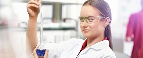 Eine junge Frau in einem Labor hält eine Pipette und betrachtet eine blaue Flüssigkeit in einem Erlenmeyerkolben mit Interesse.