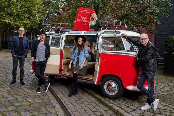 Vier Personen stehen um einen roten VW-Bus, während eine Frau auf dem Dach mit einem Schild posiert, das 'Tag der Entscheidung' zeigt.