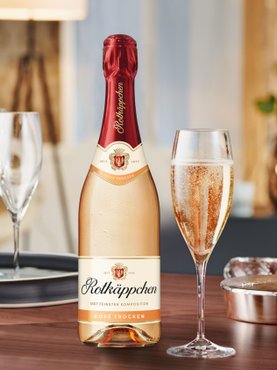 Eine Flasche Rotkäppchen Rosé Sekt steht auf einem Tisch, daneben ein Glas mit sprudelndem Sekt und eleganten Gläsern.