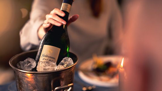 Eine Hand greift nach einer Flasche Geldermann Sekt, die in einem Eiskübel steht, während ein Kerzenlicht im Hintergrund flackert.