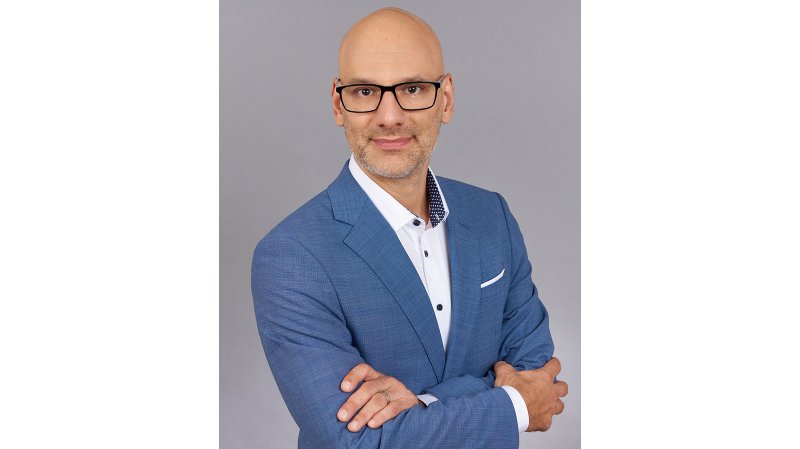 In einem eleganten blauen Anzug posiert ein Mann mit Brille, der einen professionellen und freundlichen Eindruck vermittelt.