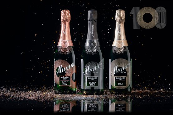 Drei Flaschen Sekt von Mumm & Co. stehen auf einem glänzenden Tisch, umgeben von glitzerndem Konfetti vor schwarzem Hintergrund.
