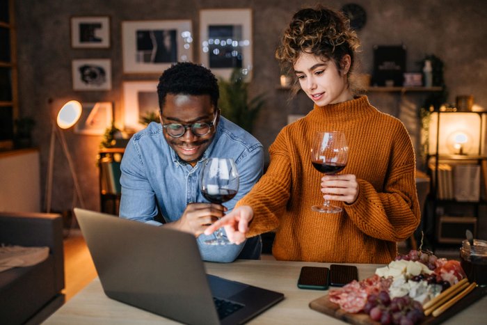 Ein fröhliches Paar genießt Rotwein, während sie auf einen Laptop schauen und sich angeregt unterhalten.