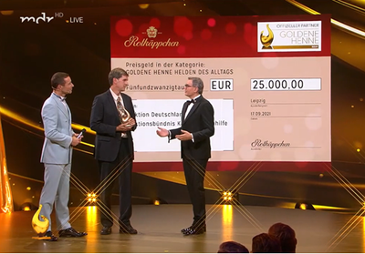 Drei Männer stehen auf einer Bühne, während einer einen Preis für die Goldene Henne entgegennimmt, umgeben von festlicher Beleuchtung.