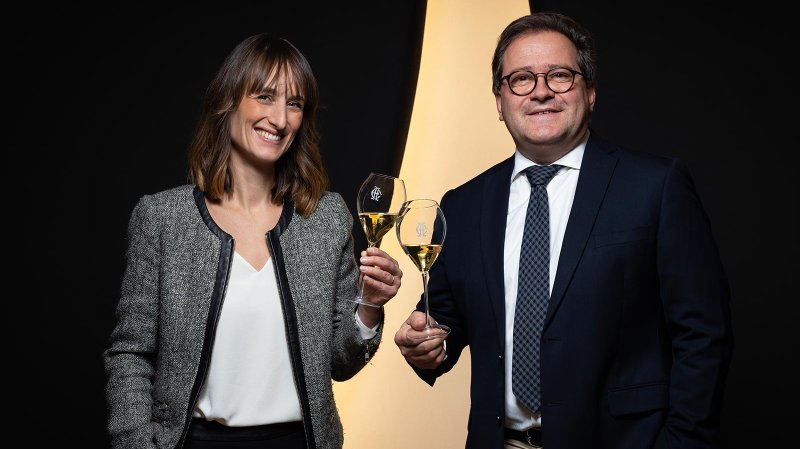 Ein Mann und eine Frau stoßen mit Gläsern voller Sekt an, beide lächeln glücklich vor einem dunklen Hintergrund.