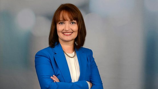 Eine lächelnde Frau mit schulterlangen braunen Haaren trägt einen blauen Blazer und steht mit verschränkten Armen vor einem unscharfen Hintergrund.