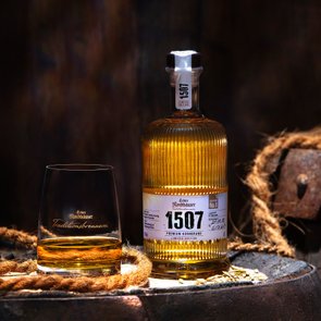 Eine Flache Echter Nordhäuser Traditionsbrennerei 1507 No. 8 und ein Glas stehen auf einem Holzfass vor dunklem, rustikalem Hintergrund.