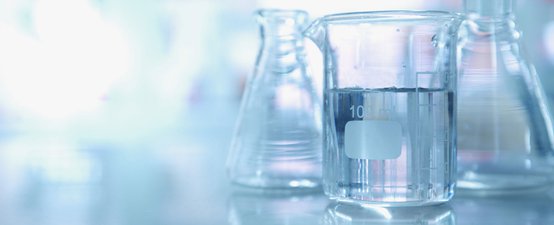 Ein Becherglas mit Wasser steht im Vordergrund, umgeben von weiteren Glasbehältern in einem hellen Laborumfeld.
