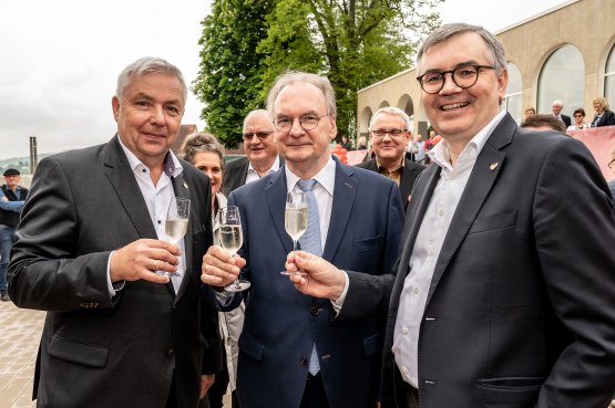 Drei Männer in Anzügen stoßen mit Sektgläsern an, während eine Gruppe von Menschen im Hintergrund lächelt und zuschaut.