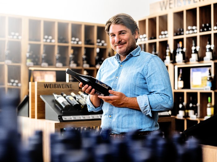 A smiling man in a wine shop holds a bottle of wine in his hand, surrounded by shelves full of wine bottles.