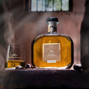 Eine edle Flasche Roggenkornbrand steht auf einem Holzfass, daneben ein Glas mit goldenem Inhalt, umgeben von Kastanien.