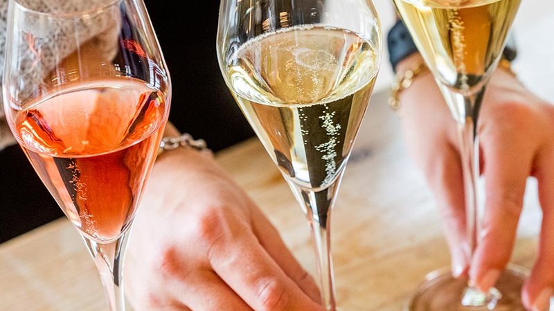 Drei Gläser mit verschiedenen Schaumweinen stehen auf einem Holztisch, während eine Hand das mittlere Glas hält.