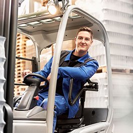 Ein junger Mann in blauer Arbeitskleidung sitzt lächelnd in einem Gabelstapler, umgeben von Holzpaletten im Hintergrund.