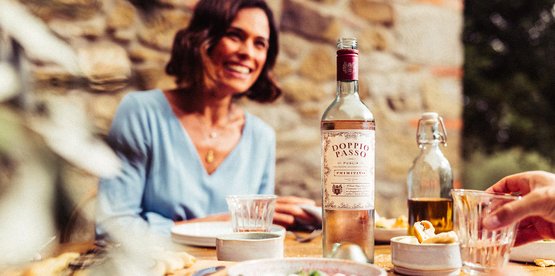Auf einem rustikalen Tisch stehen eine Flasche Doppio Passo Wein und verschiedene kleine Gerichte, während eine Frau lächelt.