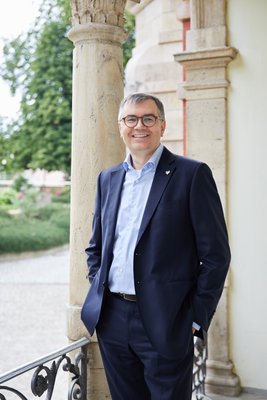 Christof Queisser in einem eleganten Anzug lächelt selbstbewusst, während er an einer Säule in einem schönen Garten steht.