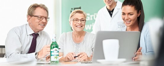 Drei Personen sitzen an einem Tisch und schauen lächelnd auf einen Laptop, während ein junger Mann im Hintergrund steht.