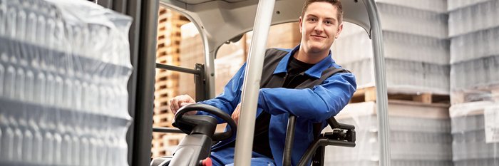 A young man in blue work clothes sits smiling in a forklift, surrounded by pallets and bottles.
