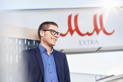 Mit einem breiten Lächeln lehnt der Mann an einer Kiste, während das Sonnenlicht hinter ihm strahlt und das MM extra Logo hervorhebt.