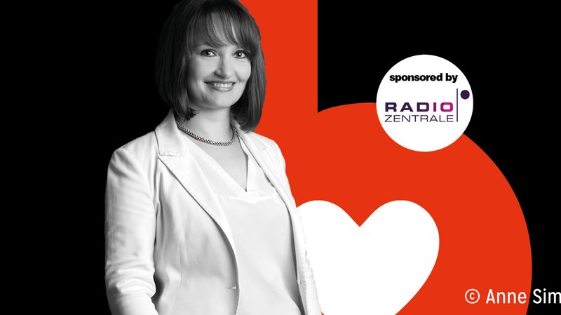 Vor einem roten Herzsymbol posiert eine Frau in einem eleganten weißen Outfit, das ihre positive Ausstrahlung unterstreicht.