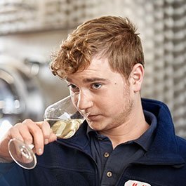 Der Mann in blauer Jacke hält ein Weinglas und betrachtet es aufmerksam, während er in einer modernen Umgebung sitzt.